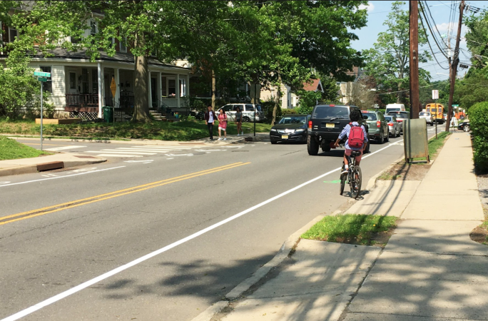 People walking down a sidewalk with a bike lane adjacent with a person riding a bike on a 2-lane street