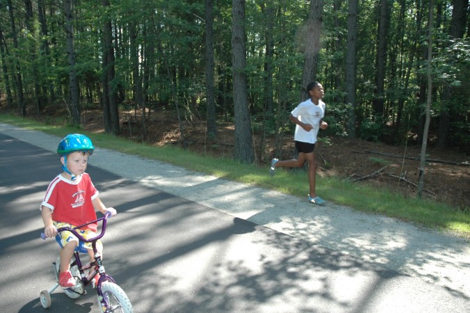 Photo of a child riding a bicycle and a runner both using a traditional greenway with a natural surface shoulder