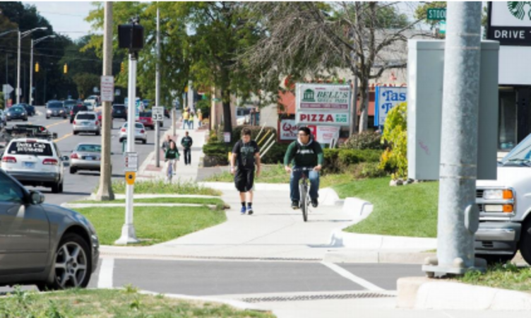 In the foreground one person walking and one person riding a bike on a multi-use sidepath with a turning vehicle at the upcoming intersection